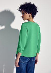 Top verde a maniche lunghe con scollatura rotonda e polsini arricciati, abbinato a jeans blu. Texture liscia, colore uniforme e vestibilità comoda.