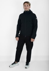 Giacca nera con cappuccio e zip frontale, dotata di tasche laterali e texture liscia. Il modello indossa pantaloni neri e sneakers di colore chiaro.