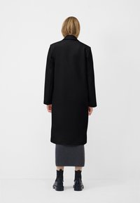 Cappotto lungo nero con un design texture, dotato di colletto e lunghezza fino al polpaccio. Abbinato a una gonna grigia e stivaletti neri alla caviglia.
