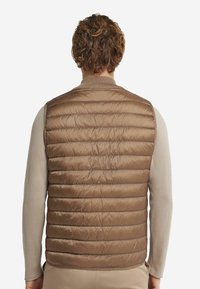 Brun pufferväst med horisontellt quiltat mönster, lättviktigt tyg och ribbad krage. Bärs över en långärmad beige skjorta.