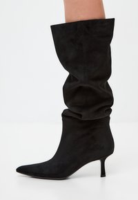 Botas de ante negras hasta la rodilla con un diseño caído, puntera afilada y un tacón delgado y bajo. Textura suave en todo.