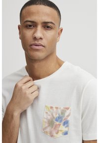 Hvid kortærmet t-shirt med en multicolor tie-dye lomme med blå, grønne og pink mønstre. Blødt stof tekstur er synligt.