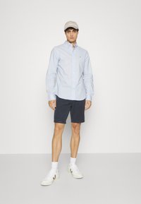 Lichtblauw overhemd met knopen, marineblauwe shorts, witte sneakers en een beige pet; zachte stof,slim fit, minimale branding op het shirt.