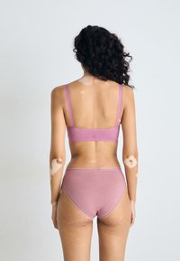 Kvinne med vitiligo på armene og ryggen iført mauve-BH og high-waist truser, som står mot en ensfarget lys grå bakgrunn, med ryggen mot kamera.