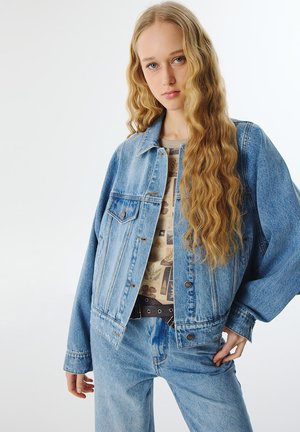 Chaqueta de mezclilla azul claro con un corte holgado, grandes bolsillos y cierre de botones; llevada sobre una camisa con estampado y combinada con jeans de mezclilla claros.