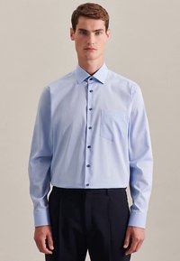 Camicia azzurro chiaro con colletto a punta, maniche lunghe e taschino sul petto. Presenta bottoni scuri e un tessuto liscio e strutturato.