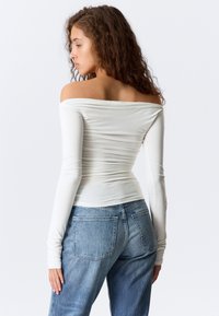 Ung kvinne med krøllete hår iført en tettsittende hvit langermet topp med bar skuldre og lyseblå denimjeans, sett fra siden.