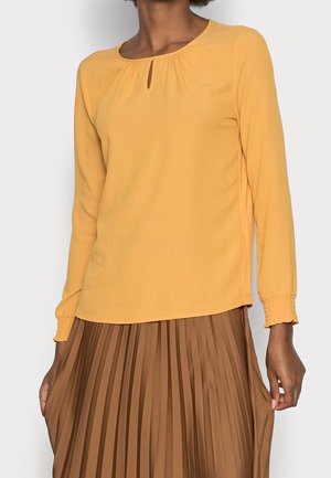 Blouse couleur moutarde à manches longues, avec un décolleté en clé et des poignets froncés. Le tissu est lisse, associé à une jupe plissée marron.