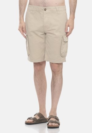 Uomo con pantaloncini cargo beige e sandali marroni a doppio cinturino, in piedi con le mani rilassate lungo i fianchi su sfondo bianco.