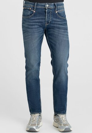 Uomo con jeans di denim blu e sneakers argentate e bianche in piedi davanti a uno sfondo chiaro e uniforme.