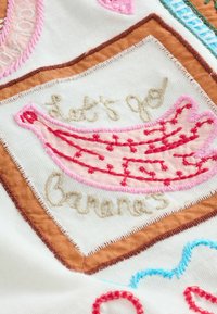 Embroidery of a pink banana with red stitched patterns on white fabric, surrounded by brown and pink embroidered text and borders.