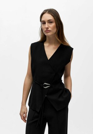 Frau trägt ärmelloses schwarzes Wickeltop mit silberner Schnallengürtel und schwarze Hose, steht vor schlichtem weißem Hintergrund.