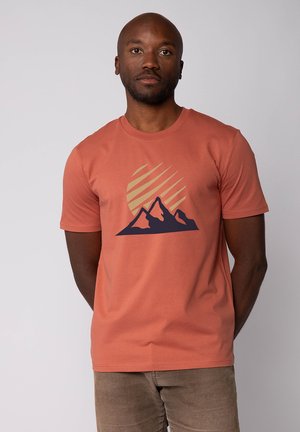 Man met een roestkleurig T-shirt met een afbeelding van marineblauwe bergen en een beige gestreepte zon op de voorkant, staand met zijn handen achter zijn rug.