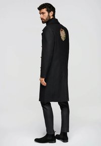 Langer schwarzer Wollmantel mit hohem Kragen, dekorativer Stickerei auf dem Rücken und goldfarbenen Knöpfen. Kombiniert mit schlanken Hosen und Stiefeln.