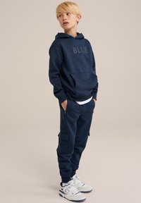 Sweat à capuche bleu marine avec le texte "BLUE", design de poche à l'avant, associé à un pantalon de survêtement assorti avec des poches cargo et des poignets resserrés. Baskets blanches.