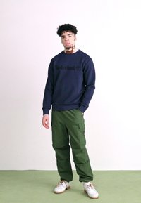Tânăr cu vitiligo stând în picioare, purtând un hanorac Timberland de culoare navy, pantaloni cargo verzi și pantofi albi într-un decor minimalist.