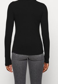 Svart ribbad långärmad topp med en åtsittande design och hög hals. Sömmen är något längre bak, ihop med grå jeans.