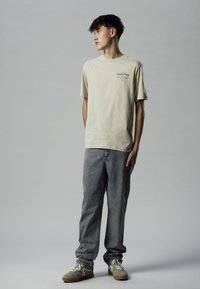 Beige katoenen t-shirt met groene tekst "Shady Pines Golf Club", gecombineerd met vervaagde grijze denim broek en lichtgekleurde sneakers.