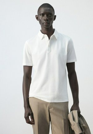 Hombre negro con polo blanco de manga corta y pantalones beige, sosteniendo una chaqueta beige sobre un fondo liso y claro.