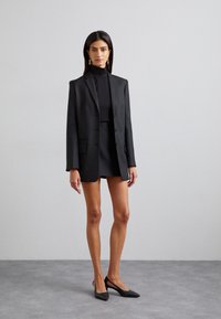 Veste noire cintrée avec une texture lisse, fermeture à un bouton, et deux poches avant, associée à une jupe mini assortie et des escarpins à nez pointu.