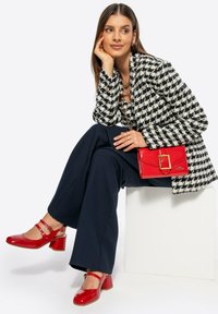 Blazer de pata de gallo en blanco y negro, pantalones de pierna ancha en azul marino, zapatos de charol rojos con tacones de bloque y un bolso rojo con herrajes dorados.