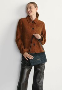 Czekoladowa zamszowa kurtka z kołnierzem i czarnymi guzikami, w połączeniu z czarną torebką o fakturze z złotym logo, zestawiona z czarnymi spodniami.