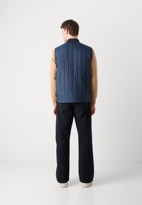 Colete acolchoado azul-marinho com gola alta, usado sobre uma camisa de manga longa bege. Combinado com calças escuras de perna larga e ténis claros.