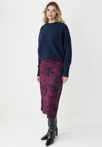 Marineblauer, strukturierter Pullover kombiniert mit einem bordeauxroten, floralen Midirock. Das Modell trägt kniehohe schwarze Stiefel, die ein geschichtetes Design und Farben zeigen.