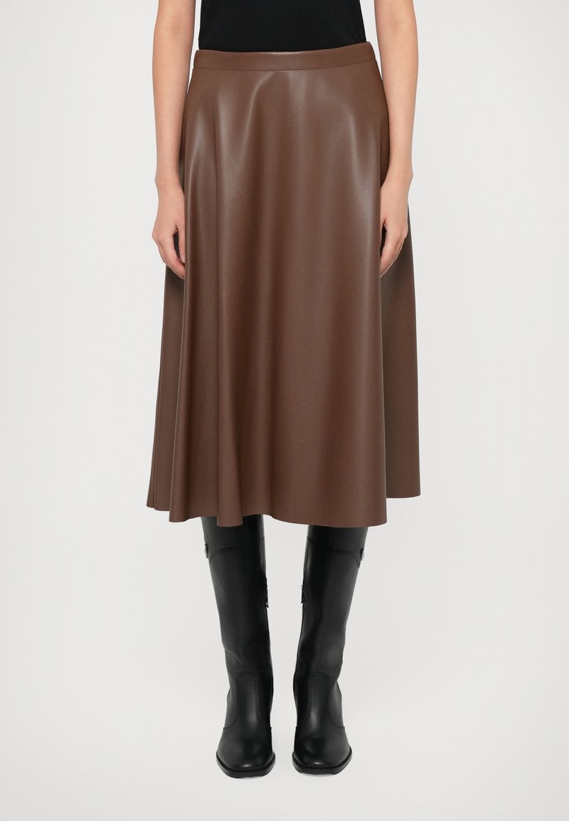 Jupe en cuir marron de longueur midi portée avec des bottes hautes noires et un haut noir, visible de la taille aux pieds sur un fond blanc.
