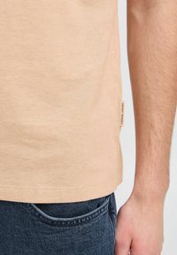 Light peach t-shirt in soft, textured cotton, featuring a straight hem and a small label on the side. Worn with dark denim jeans.