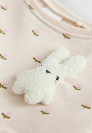 Gaiši rozā džempera ar ziedu izšuvumiem, kurā ir pūkaina balta truša aplikācija ar melnām izšūtām acīm un teksturētu apdari.