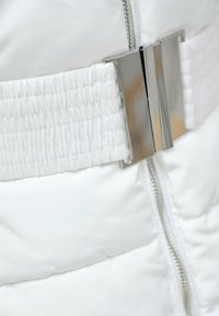 Chaqueta acolchada blanca con un cinturón de tela fruncido y una hebilla de metal plateado, y un cierre de cremallera lateral. Textura suave y diseño minimalista.