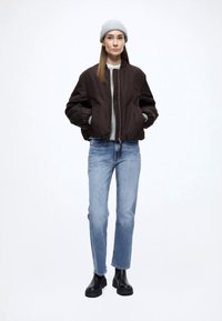 Hnedá bomber bunda, sivý sveter, svetlomodré rifle a čierne členkové čižmy. Modelka má na hlave sivú pletenú čiapku a dlhé vlasy.