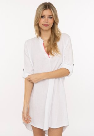 Junge Frau mit langen blonden Haaren, die ein weißes, langärmeliges Hemdblusenkleid mit hochgekrempelten Ärmeln und einem leicht rosa Halsausschnitt trägt.