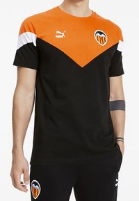 Persona con una camiseta deportiva negra y naranja de Puma y pantalones negros, ambos con el logo del club de fútbol Valencia.