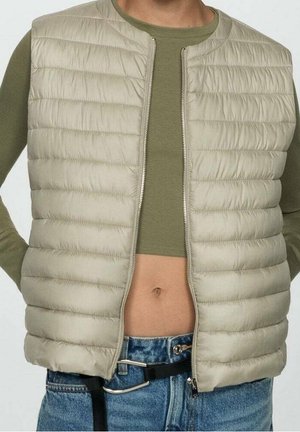 Beige quilted sleeveless vest worn open over olive green long sleeve crop top and blue denim jeans with black belt.