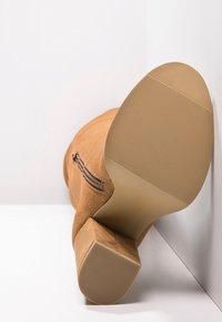 Beige mockasinhögboot med blockklack, dragkedjelement och en texturerad sula för bra grepp, placerad mot en vit bakgrund.