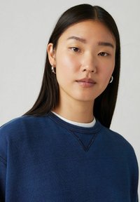 Marinblå sweatshirt med rundad halsringning, strukturerad tyg och kontrasterande sömmar. Modellen bär ett vitt underplagg och hoopörhängen.