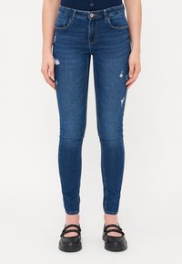 Vékony kék farmernadrág, kopott részletekkel, standard öt zsebes dizájnnal és közepes derékvonallal. Fekete slip-on cipővel viselve.