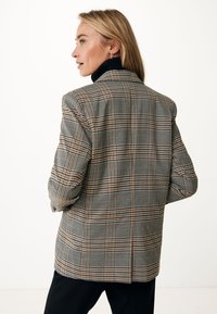 Blazer a quadri nei toni del marrone, beige e blu, con spalle strutturate, vestibilità su misura e tessuto testurizzato. Nella descrizione è presente un dolcevita nero indossato sotto.