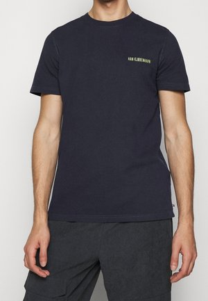 Mężczyzna ubrany w ciemnogranatowy, krótki rękaw t-shirt z małym napisem "HAN KJØBENHAVN" na lewej piersi oraz ciemne spodnie typu cargo.