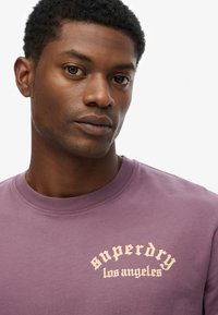 Camiseta de crewneck morada hecha de algodón con una textura suave. Presenta la inscripción "superdry los ángeles" impresa en un font arqueado de color beige claro.