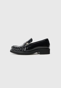 Mocassins pretos em cabedal brilhante com uma borda decorativa franzida, sola de borracha plana e um pequeno logotipo metálico em destaque na faixa.