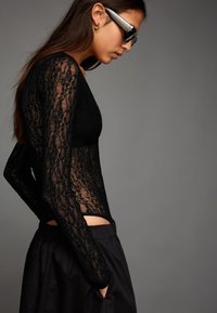 Žena v čiernom čipkovanom topu s dlhými rukávmi a čiernych nohaviciach, s dlhými hnedými vlasmi, veľkými slnečnými okuliarmi a zlatým kruhovým náušníkom na sivom pozadí.