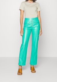 Woman wearing beige short-sleeve top and bright turquoise high-waist leather pants with front zipper and brown open-toe heeled sandals.