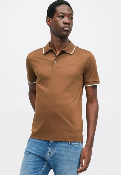 Camisa polo marrón con ribete y botones blancos, con mangas cortas y un diseño ajustado, combinada con jeans azules.