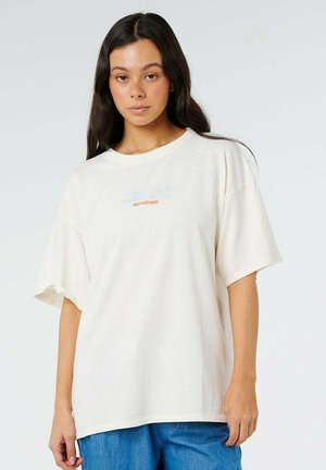 Jonge vrouw met lang donker haar, die een oversized wit T-shirt met kleine bedrukte tekst draagt en een blauwe spijkerbroek, staand tegen een effen achtergrond.