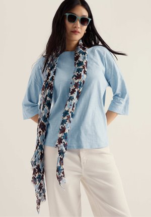 Mujer con gafas de sol color verde azulado, una blusa azul claro de tres cuartos de manga, pantalones crema y una larga bufanda floral con flores azules y marrones.