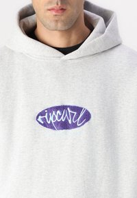 Szara bluza z kapturem wykonana z miękkiego materiału. Na przodzie znajduje się fioletowo-niebieskie haftowane logo. Prosty design o luźnym kroju.