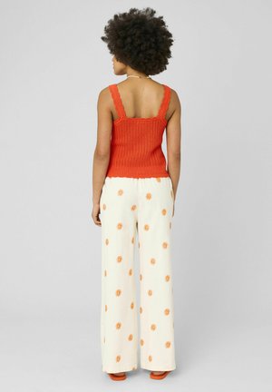 Femme aux cheveux bouclés portant un débardeur tricoté orange, un pantalon blanc à jambes larges avec des motifs de soleil orange, et des sandales orange, vue de dos.